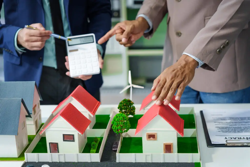 A meeting at an office desk focuses on teamwork with a house model and discussions on electric cars